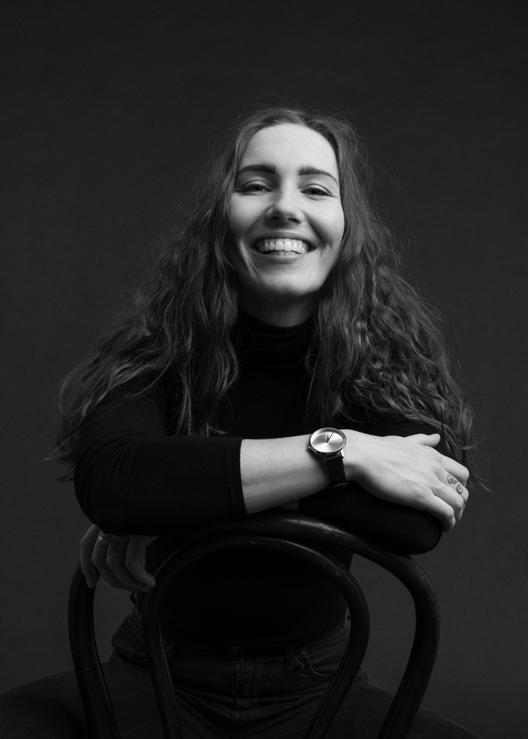 A woman with long wavy hair and a watch on her wrist sits on a chair backward, as if pausing after a cycle course, smiling broadly at the camera against a plain dark background. She is wearing a long-sleeve, high-neck black top.