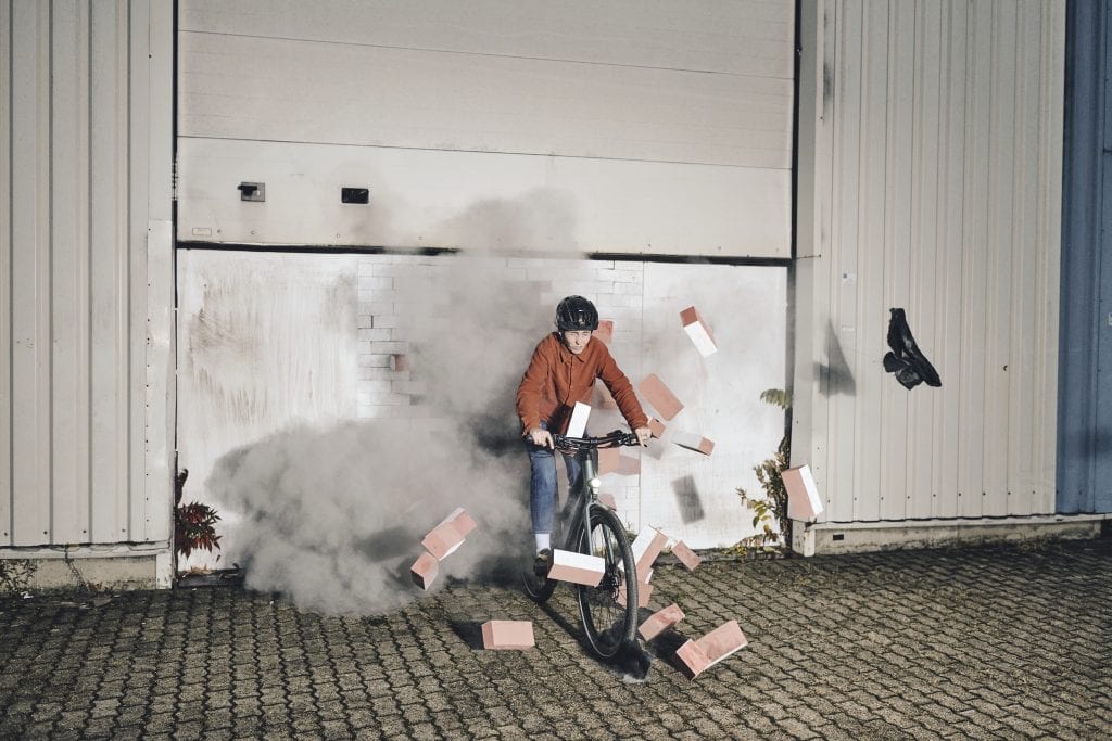 Eine Person mit Helm fährt mit einem Mountainbike durch eine Wand. Rauch und Ziegel fliegen umher, ein Schuh löst sich. Die Szene spielt sich draußen auf einem gepflasterten Boden ab.