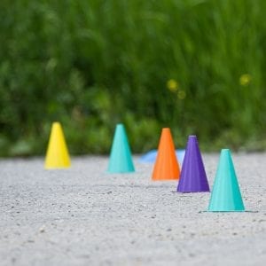 Fünf bunte Plastikkegel – gelb, grün, orange, violett und blau – stehen in einer Reihe auf einer Kiesfläche im Freien, perfekt für Mountainbike- oder Fahrradtrainingsübungen, mit grünem Gras im unscharfen Hintergrund.