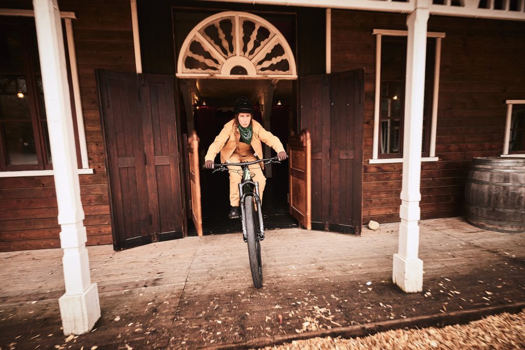 Eine Person mit Helm und hellbrauner Kleidung fährt mit einem Mountainbike aus der Doppeltür eines Holzgebäudes mit einem dekorativen Fächerfenster über dem Eingang.