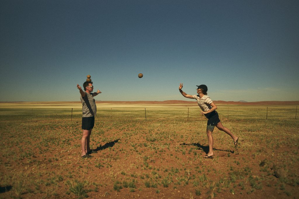 Zwei Personen spielen auf einem weiten, offenen Feld unter strahlend blauem Himmel Fangen. Einer balanciert zwei Bälle auf dem Kopf und fängt gleichzeitig einen dritten, während der andere, barfuß und mit Helm, bei jedem Wurf einer unvorhersehbaren Richtung folgt.