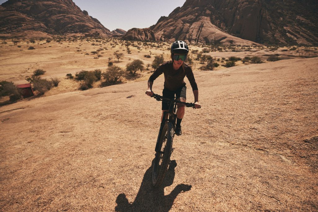 Eine Person mit Helm und Sonnenbrille fährt mit einem Mountainbike einen felsigen, sonnenbeschienenen Hang hinauf und bewältigt dabei eine anspruchsvolle Strecke durch eine trockene, wüstenähnliche Landschaft mit schroffen Hügeln im Hintergrund.
