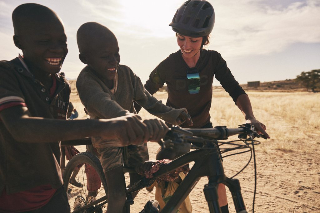 Drei lächelnde Kinder und ein Erwachsener versammeln sich an einem sonnigen Tag im Freien auf einer trockenen Wiese um ein Fahrrad. Der Erwachsene trägt einen Helm, was auf eine Radtour oder einen Radkurs hindeutet. Die Stimmung ist fröhlich und verspielt.
