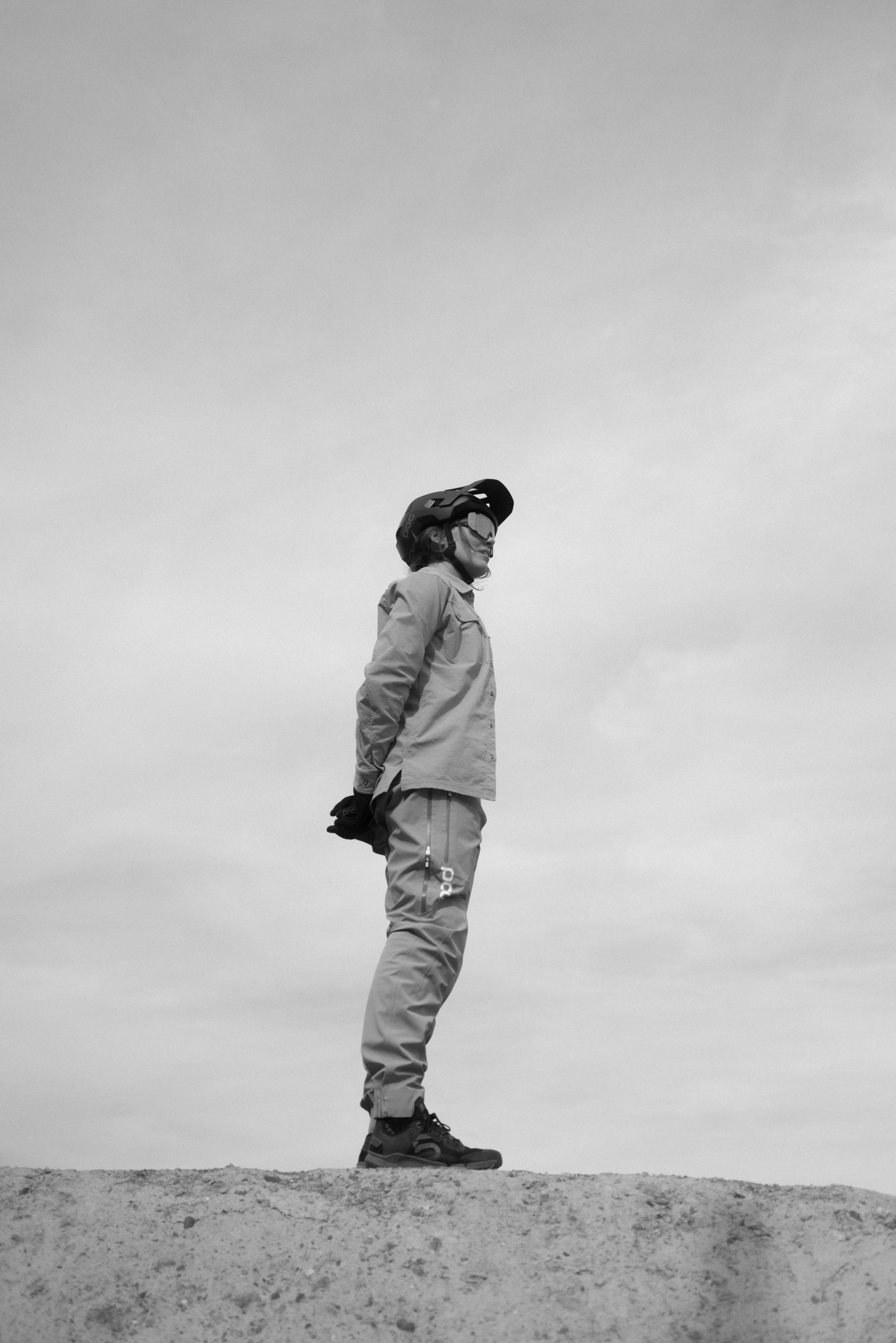 Auf einem Schwarzweißfoto steht eine Person in Uniform, mit Helm und Handschuhen auf einer ebenen Fläche, die Hände auf dem Rücken verschränkt, und blickt während einer Radtour zur Seite vor einem bewölkten Himmel.