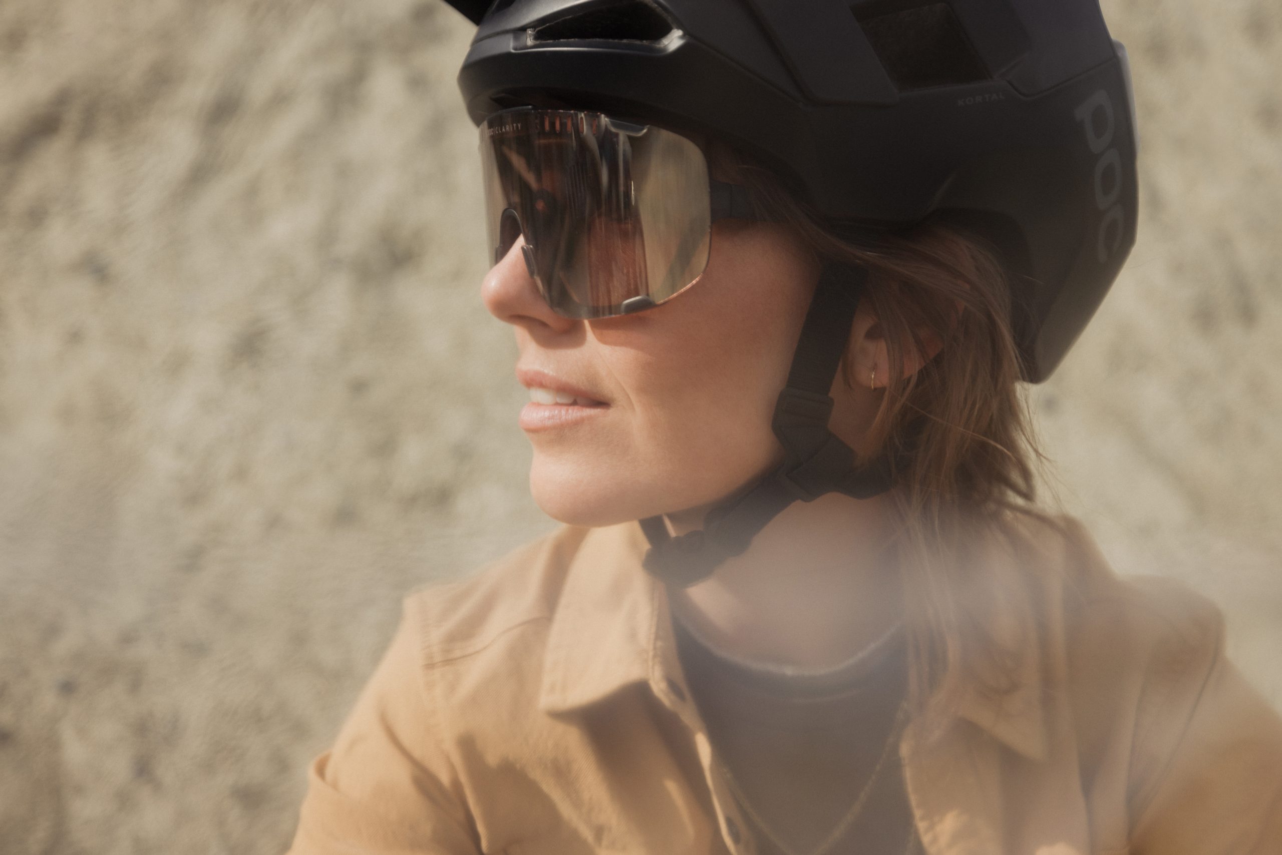 Ein Model mit schwarzem Helm und großer Sonnenbrille blickt zur Seite und lächelt sanft. Der Hintergrund ist unscharf, die Person trägt eine hellbraune Jacke und ist bereit für ein Mountainbike-Abenteuer.