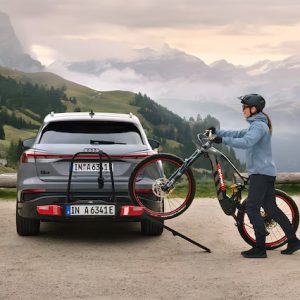 Eine Person mit Helm und Outdoor-Bekleidung lädt ein Mountainbike auf die Ladefläche eines grauen Audi SUV, des neuesten Modells, das in einer malerischen Berglandschaft mit nebligen Hügeln und Grün im Hintergrund geparkt ist.