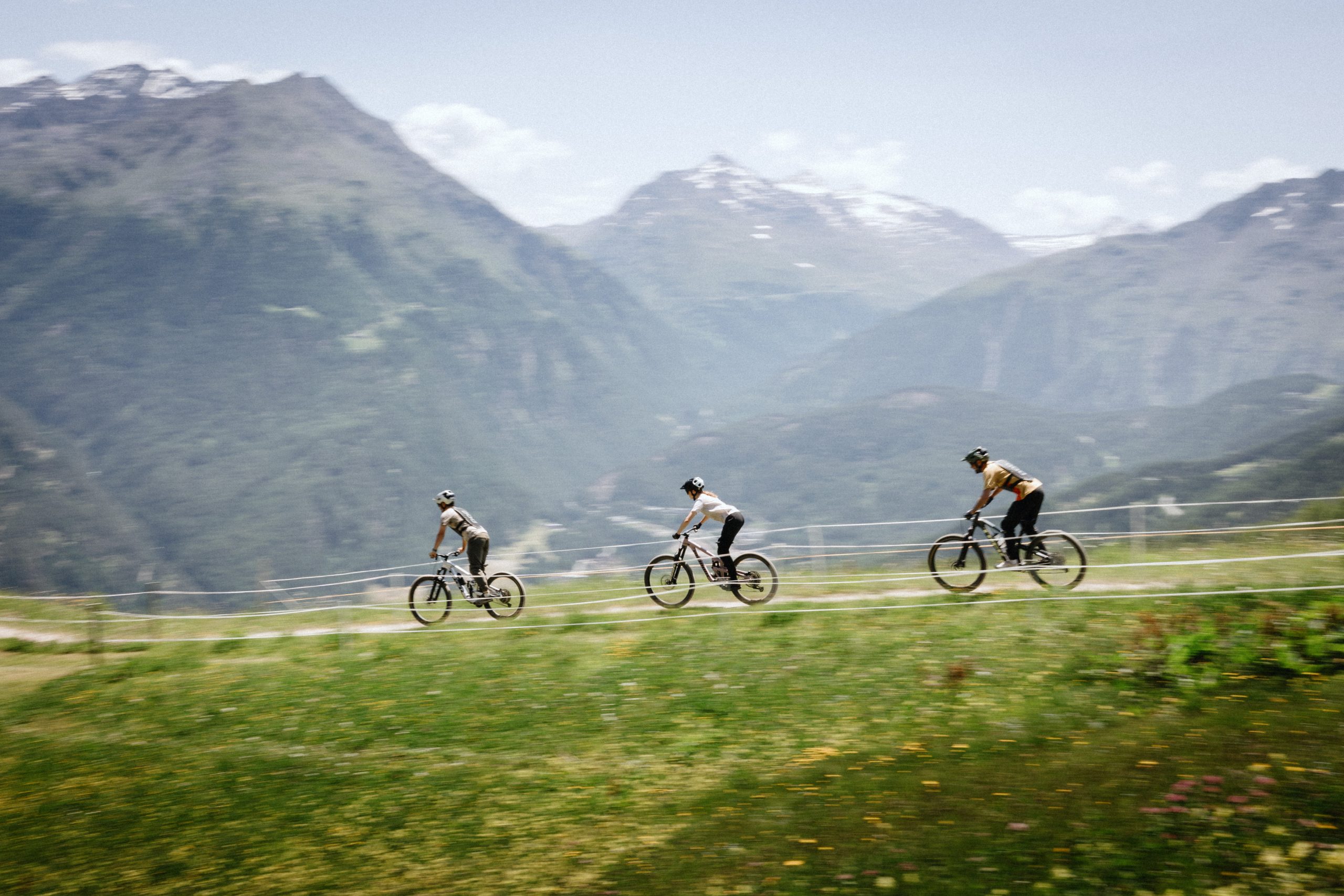 Drei Radfahrer fahren auf einem gewundenen Bergpfad mit schneebedeckten Gipfeln und grünen Tälern im Hintergrund unter einem teilweise bewölkten Himmel. Die Szene suggeriert Abenteuer im Freien und eine alpine Landschaft.