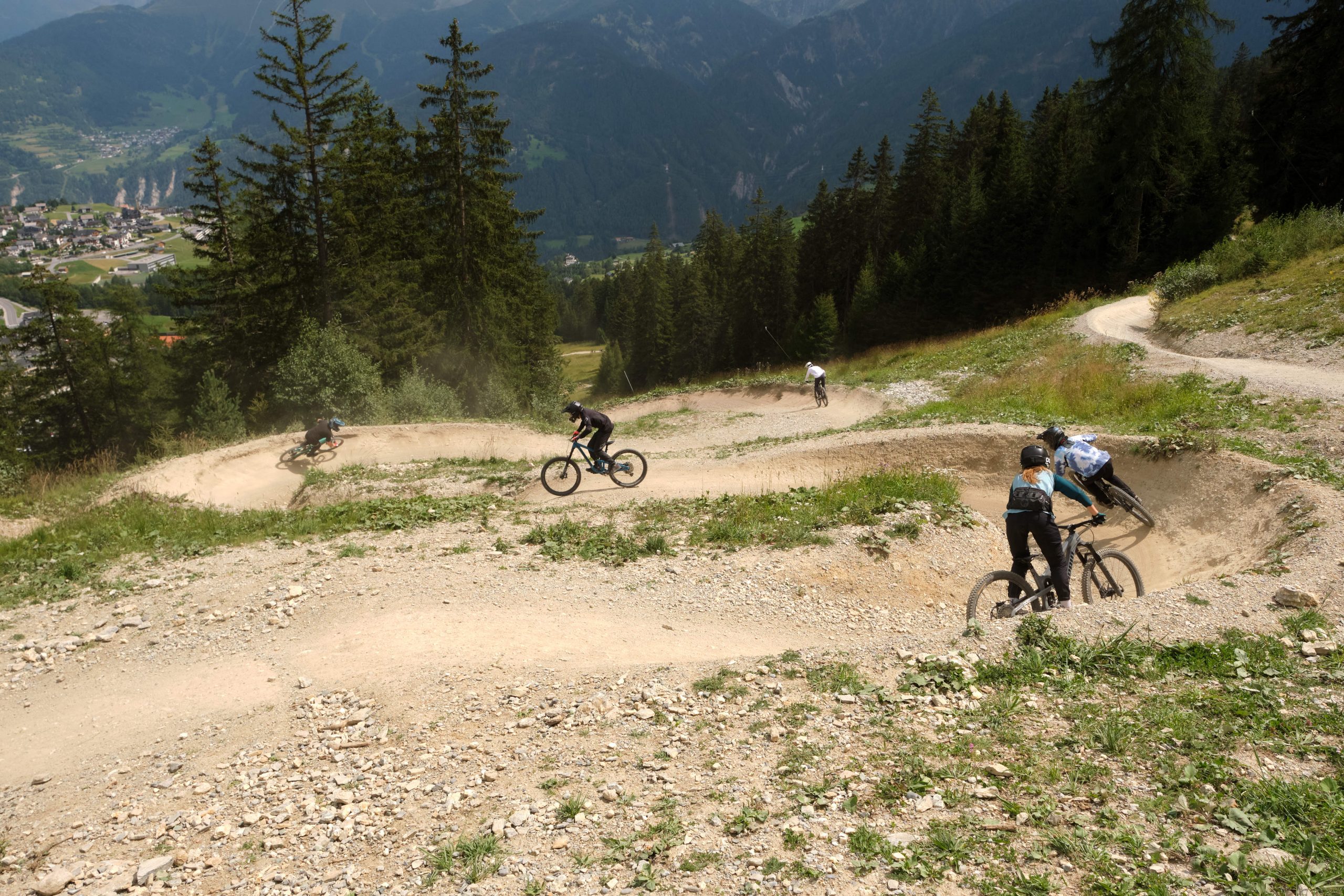 Vier Fahrer rasen über einen gewundenen Schotterweg beim 13.-15.6. | USI | MTB Downhill Camp in Serfaus Fiss Ladis, durch scharfe Kurven und üppiges Grün zwischen hohen Bäumen, mit Bergen und einem Tal im Hintergrund.