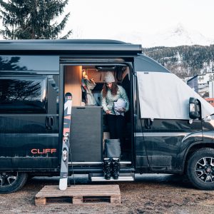 Eine Frau in Winterkleidung steht an der Tür eines schwarzen Wohnmobils auf dem 23.-25.1. SUNLIGHT Winter Camp & Ride in Sölden, Österreich. Sie hält einen Becher in der Hand, Skier in der Nähe und verschneite Berge im Hintergrund. Ski