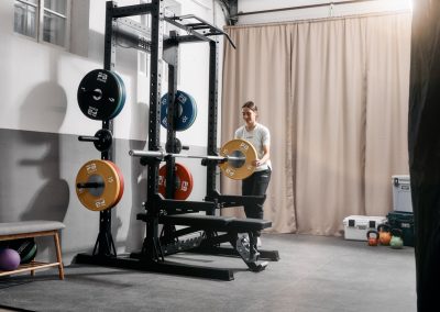 Eine Person in Sportkleidung lädt eine gelbe Hantelscheibe auf eine Langhantel in einem gut beleuchteten Fitnessstudio, in dessen Nähe eine Kniebeugenablage, eine Bank und bunte Hantelscheiben zu sehen sind.