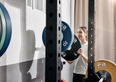 Eine Frau in einem weißen Hemd legt eine Hantelscheibe mit der Aufschrift "PB Strong" auf eine Langhantel in einem Fitnessstudio, umgeben von anderen bunten Hantelscheiben und Trainingsgeräten.