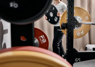 Eine Person in einem Fitnessstudio lädt oder entlädt Gewichtsplatten auf eine Langhantel in einem Squat Rack. Mehrere bunte Hantelscheiben sind im Vorder- und Hintergrund zu sehen. Das Gesicht der Person ist nicht zu sehen.