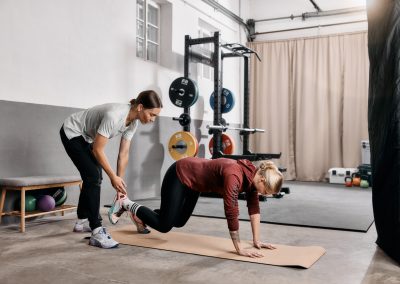Ein Trainer hilft einer Frau beim Bergsteigen auf einer Matte in einem Fitnessstudio, während im Hintergrund Gewichthebegeräte und Kettlebells zu sehen sind.