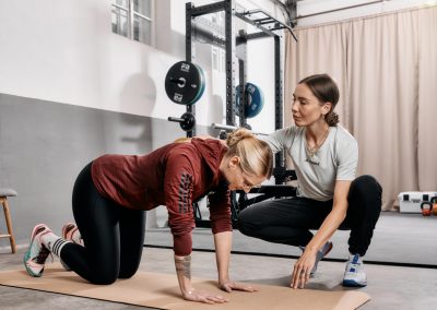 Eine Frau kniet auf einer Yogamatte und macht eine Übung, während ein Trainer neben ihr hockt und sie in einem Fitnessstudio mit Fitnessgeräten im Hintergrund anleitet.