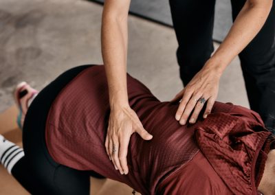 Eine Person, die eine kastanienbraune Jacke trägt, befindet sich in einer Dehnungs- oder Trainingshaltung auf einer Matte, während eine andere Person in einem hellen Hemd ihre Hände sanft auf den Rücken der Person legt, um ihr möglicherweise bei der Dehnung oder Haltung zu helfen.