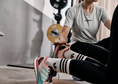 Eine Person, die ein hellgraues Terrex-Shirt trägt, hilft einer anderen Person beim Dehnen. Der Schwerpunkt liegt auf den Händen, die die Füße berühren, die bunte Turnschuhe und Adidas-Socken tragen, in einem Fitnessstudio mit Gewichten im Hintergrund.