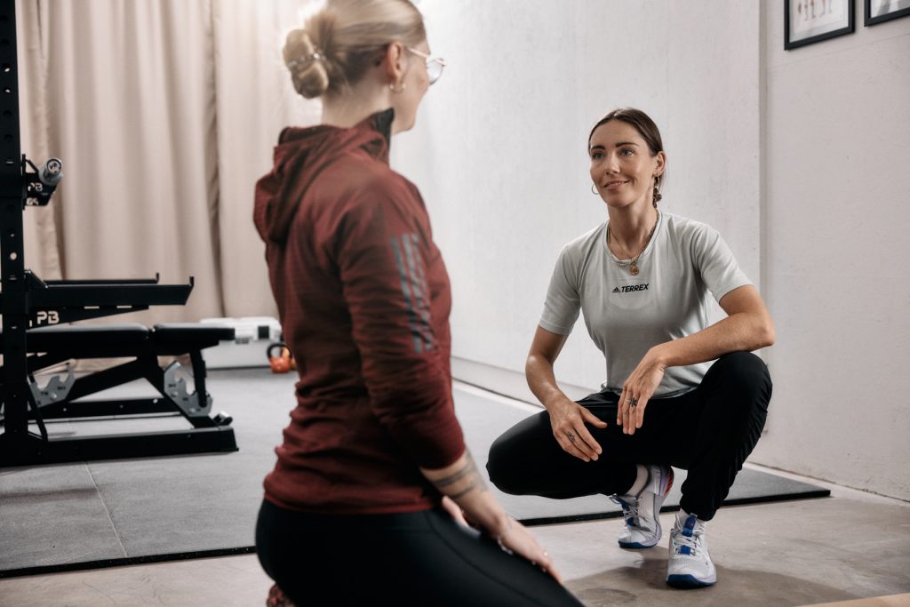 Zwei Frauen befinden sich in einem Fitnessstudio; eine kniet und spricht, während die andere, bekleidet mit einem kastanienbraunen Kapuzenpullover, aufmerksam zuhört. Im Hintergrund sind Fitnessgeräte und eine Bank zu sehen.