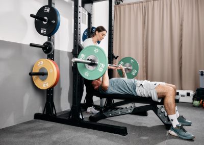 Ein Mann liegt auf einer Bank und drückt eine Langhantel mit Gewichten, während eine Frau hinter ihm steht und ihm dabei hilft. Im Hintergrund sind Fitnessgeräte und Hantelscheiben zu sehen.