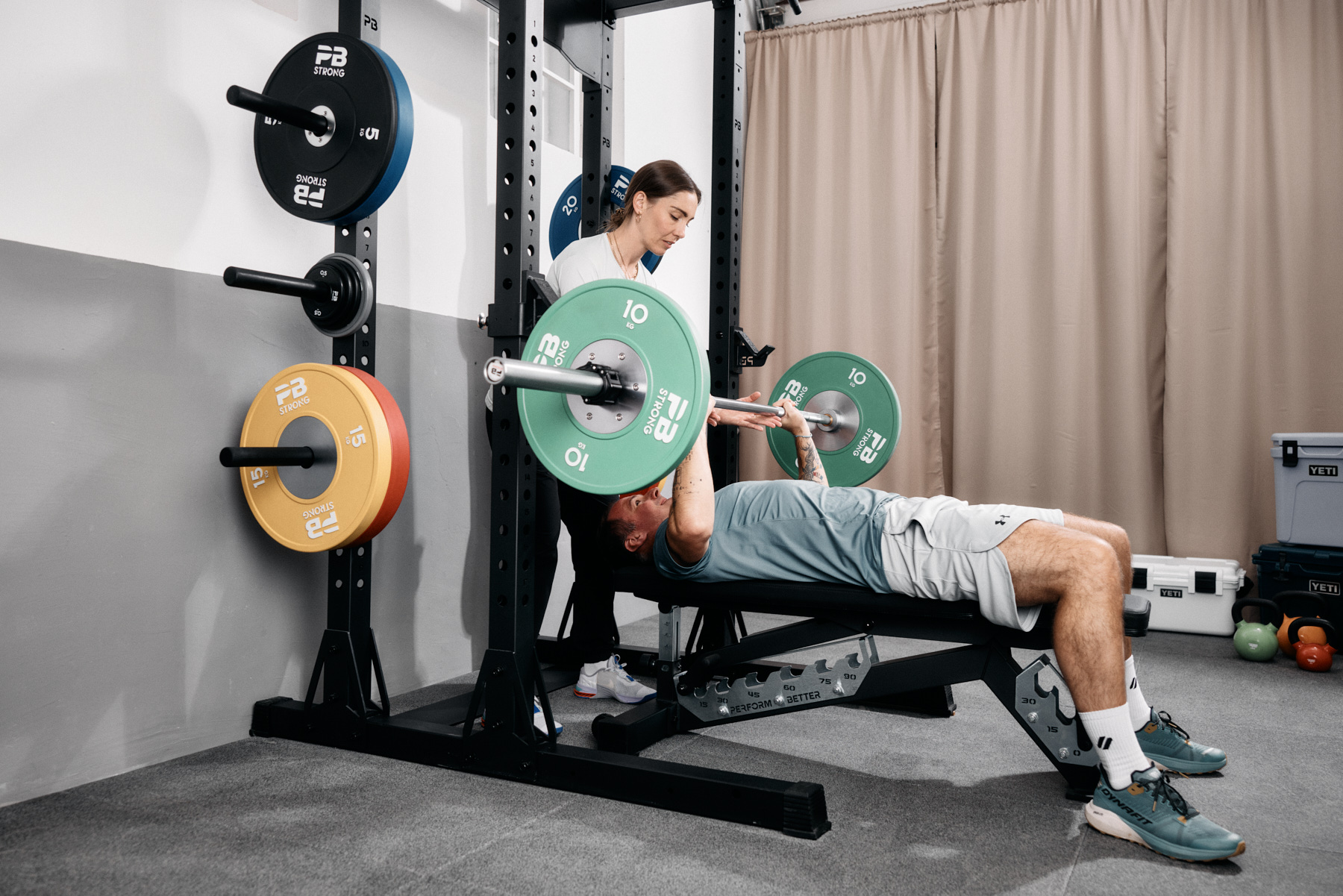 Ein Mann liegt auf einer Bank und drückt eine Langhantel mit Gewichten, während eine Frau hinter ihm steht und ihm dabei hilft. Im Hintergrund sind Fitnessgeräte und Hantelscheiben zu sehen.