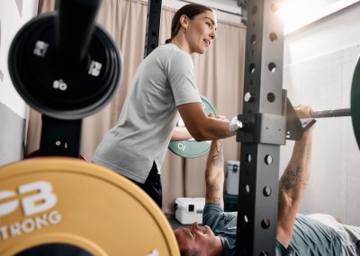 Eine Frau steht neben einem Mann, der auf einer Bank liegt und ihn beobachtet, während er in einem Fitnessstudio eine Langhantel mit Gewichten hebt. Im Vordergrund sind Hantelscheiben und Fitnessgeräte zu sehen.