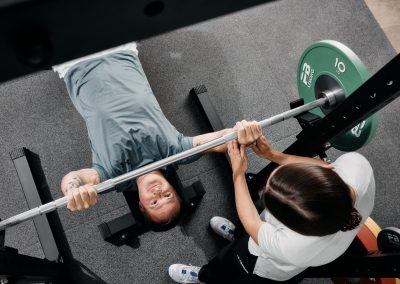 Eine Person liegt auf einer Bank und hält eine Langhantel mit grünen Gewichten, während eine andere Person neben ihr steht und ihr hilft oder sie beobachtet. Die Ansicht ist von oben und zeigt sowohl die Personen als auch die Fitnessgeräte.