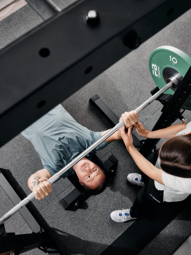 Eine Person liegt auf einer Bank und hält eine Langhantel mit Gewichten, während eine andere Person daneben steht, um zu helfen und Unterstützung anzubieten. Das Bild ist von oben aufgenommen und zeigt den Heber, der nach oben schaut und lächelt.