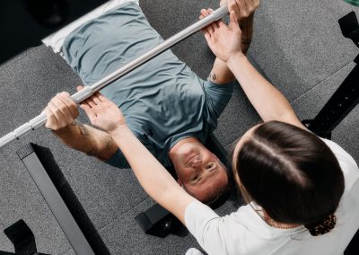 Eine Person liegt auf einer Bank und hält eine Langhantel, während eine andere Person in der Nähe steht und sie beim Gewichtheben im Fitnessstudio unterstützt und beobachtet.