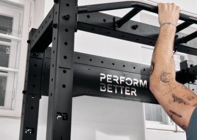 Eine Person mit Tätowierungen auf dem Arm hält den Griff eines schwarzen Fitnessgeräts mit der Aufschrift PERFORM BETTER und bereitet sich auf einen Klimmzug in einem Raum mit weißen Wänden und einem Fenster vor.