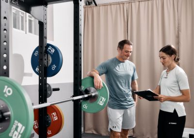 Ein Mann in Sportkleidung steht neben einer belasteten Langhantel in einem Fitnessstudio, lächelt und spricht mit einer Frau, die ein Klemmbrett in der Hand hält und offenbar eine Trainerin ist. Sie stehen neben Fitnessgeräten und Hantelscheiben.