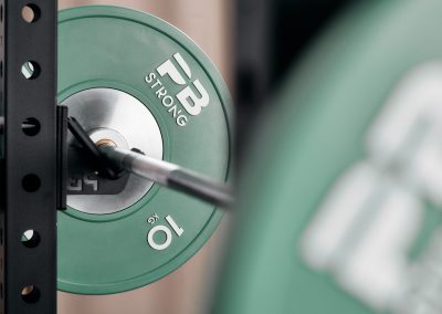 Nahaufnahme einer Langhantel mit einer grünen 10-kg-Hantelscheibe mit der Aufschrift FB STRONG, die auf einer Ablage in einem Fitnessstudio liegt, mit unscharfen Geräten im Vordergrund.