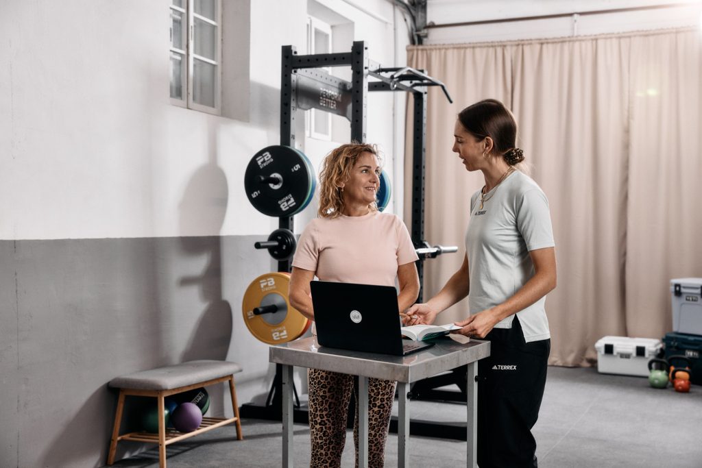 Zwei Frauen stehen in einem Fitnessstudio an einem Tisch mit einem Laptop und einem Notizbuch, unterhalten sich und lächeln. Hinter ihnen stehen Hantelscheiben, Trainingsgeräte und eine Bank mit Medizinbällen.