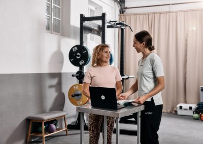 Zwei Frauen stehen in einem Fitnessstudio an einem Tisch mit einem Laptop und einem Notizbuch, unterhalten sich und lächeln. Hinter ihnen stehen Hantelscheiben, Trainingsgeräte und eine Bank mit Medizinbällen.