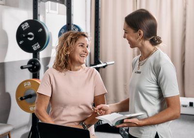 Zwei Frauen in einem Fitnessstudio lächeln und unterhalten sich an einem Tisch mit einem Laptop und einem offenen Buch. Im Hintergrund sind Geräte zum Gewichtheben zu sehen. Eine Frau gestikuliert mit ihrer Hand, während sie sich unterhalten.