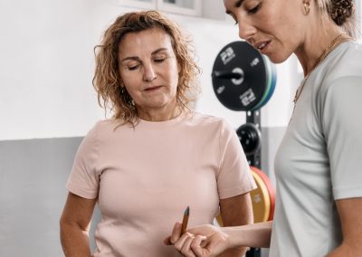 Zwei Frauen stehen in einer Turnhalle in der Nähe von Gewichthebern. Die eine hält ein Buch in der Hand und deutet darauf, während die andere mit einem Stift in der Hand und einem Klemmbrett auf dem Tisch vor ihr aufmerksam zuhört.