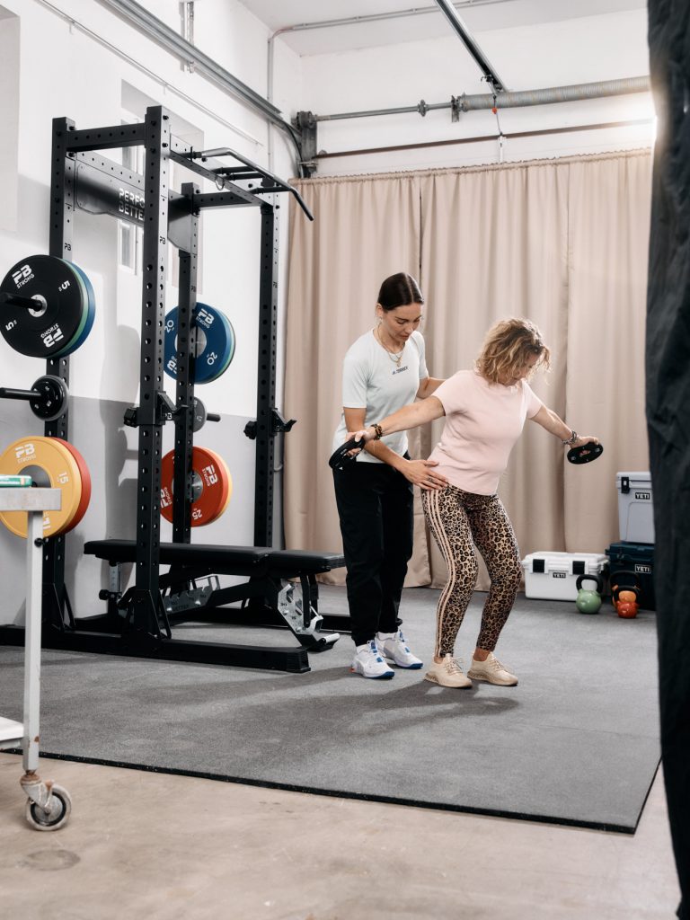 Ein Fitnesstrainer hilft einer älteren Frau bei einer Hantelübung in einem Fitnessstudio, die in der Nähe von Hantelscheiben und einer Bank steht, während im Hintergrund Trainingsgeräte zu sehen sind.