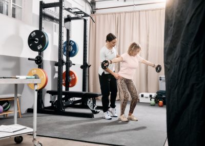 Eine Frau in Trainingskleidung hebt kleine Hanteln, während ein Trainer hinter ihr steht und ihre Form in einem Fitnessstudio mit Gewichten und Trainingsgeräten im Hintergrund anleitet.
