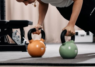 Eine Frau trainiert in einem Fitnessstudio und macht einen Liegestütz, wobei sie mit den Händen eine orangefarbene und eine grüne Kettlebell auf einer Matte festhält. Sie trägt ein weißes Hemd und eine schwarze Hose.