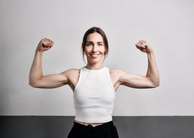 Eine Frau in einem weißen ärmellosen Oberteil lächelt selbstbewusst, während sie ihren Bizeps an einer schlichten hell- und dunkelgrauen Wand anspannt.