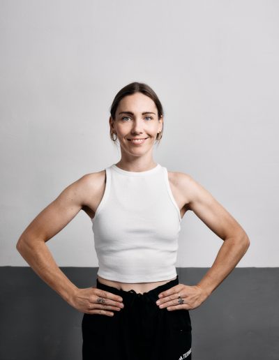 Eine Frau mit heller Haut und braunem Haar steht selbstbewusst mit den Händen in den Hüften, trägt ein weißes ärmelloses Crop-Top und eine schwarze Hose und lächelt in die Kamera vor einem schlichten grauen und weißen Hintergrund.