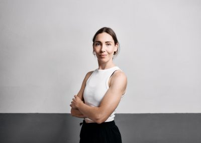 Eine selbstbewusste Frau mit hellbraunem Haar steht mit verschränkten Armen und einem leichten Lächeln vor einer zweifarbigen grauen Wand. Sie trägt ein ärmelloses weißes Oberteil und eine schwarze Hose.