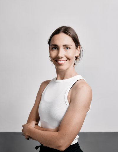 Eine Frau mit heller Haut und braunem, nach hinten gebundenem Haar lächelt in die Kamera. Sie trägt ein weißes ärmelloses Oberteil und Ohrringe und steht mit verschränkten Armen vor einem schlichten hellen Hintergrund.