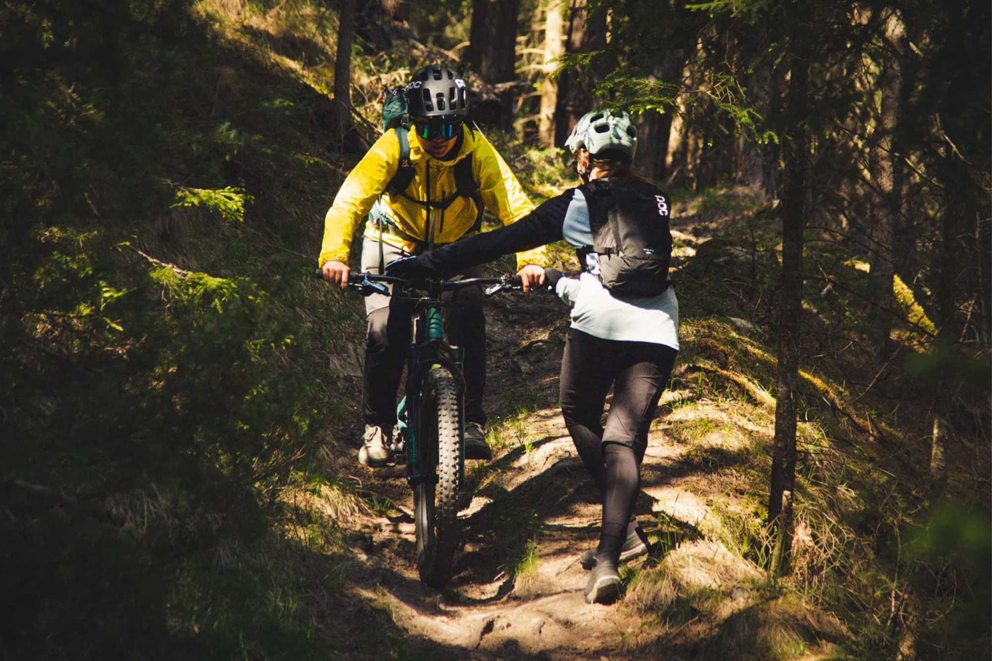 Zwei Personen mit Helmen und Rucksäcken fahren mit ihren Fahrrädern durch einen sonnenbeschienenen Waldweg, umgeben von Bäumen und Grün. Eine Person blickt nach vorne, während die andere ihnen eine Hand entgegenstreckt.