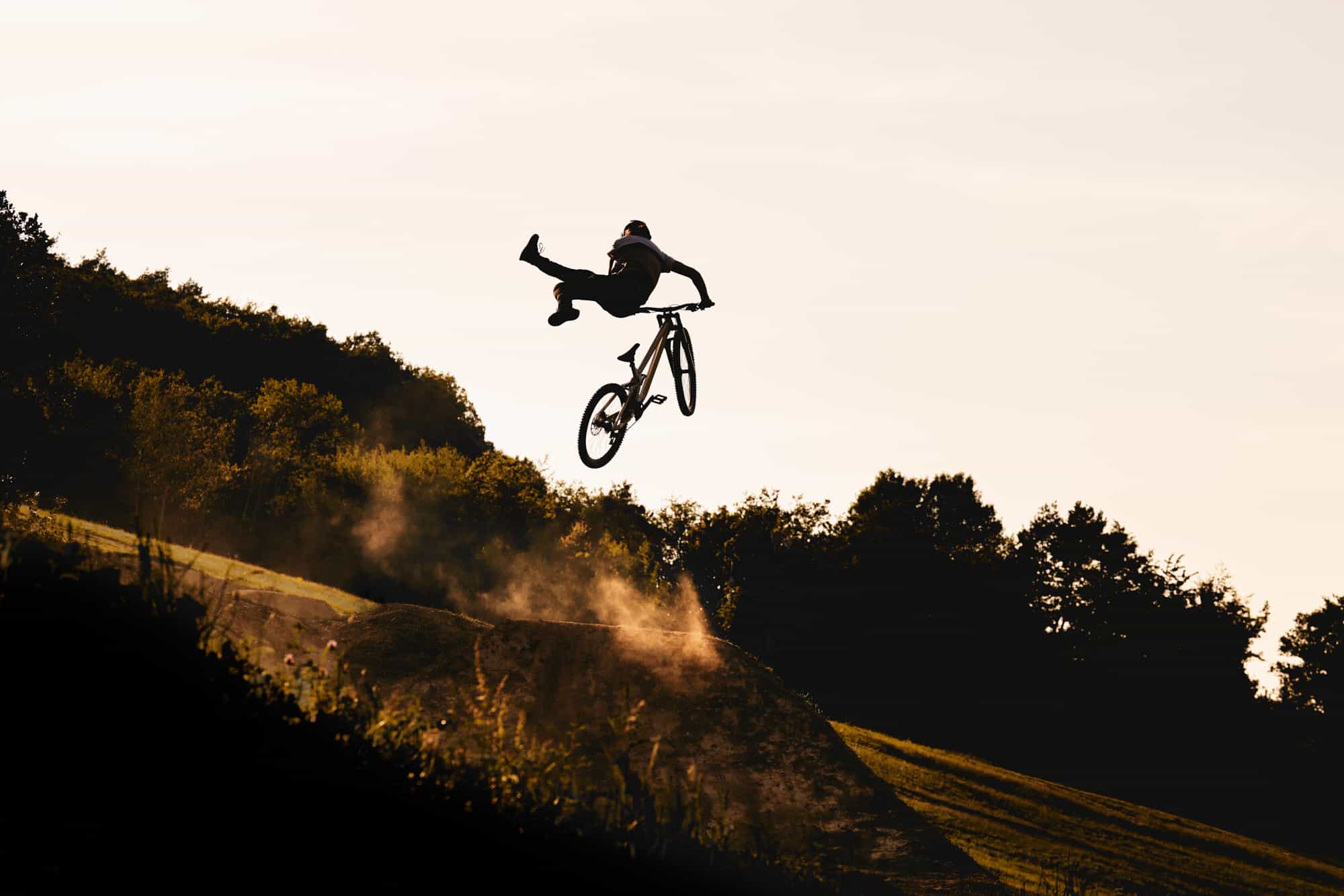 Zwei Mountainbiker mit Helm und Ausrüstung fahren auf einem steinigen, bergauf führenden Waldweg; einer fährt auf einen großen Felsen, während der andere in der Nähe steht und zwischen Bäumen und unwegsamem Gelände Unterstützung oder Führung bietet.